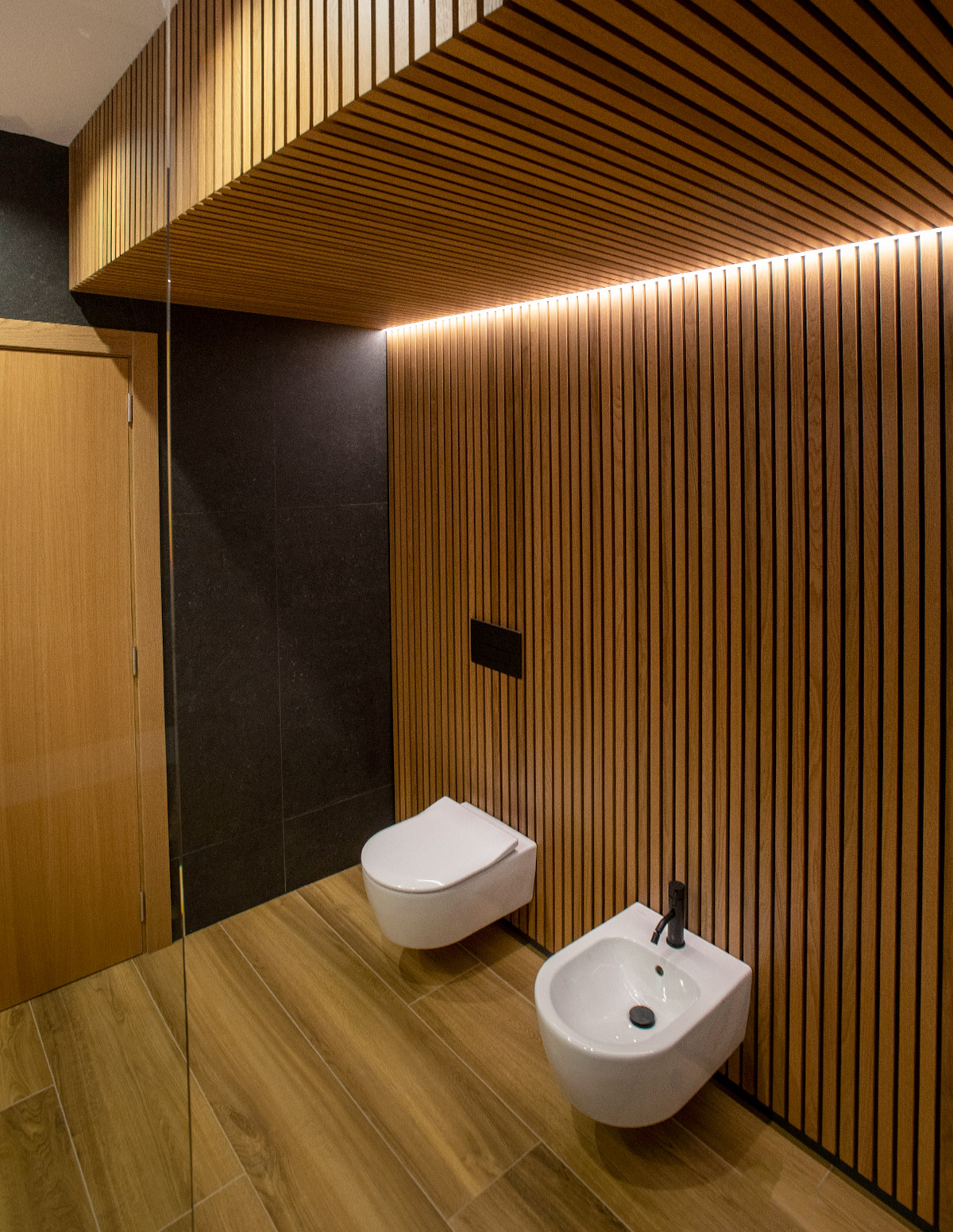 Vista de baño con sanitarios sobre madeira rastrelada e luz indirecta